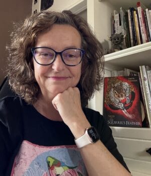 Lisette Niemand sitting in her studio with her book “SASSI and the Squirrel’s Feather” on the shelf behind her. She wears a TeeMill t-shirt featuring her own surface pattern design.