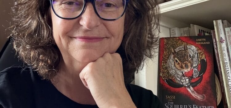 Lisette Niemand sitting in her studio with her book “SASSI and the Squirrel’s Feather” on the shelf behind her. She wears a TeeMill t-shirt featuring her own surface pattern design.
