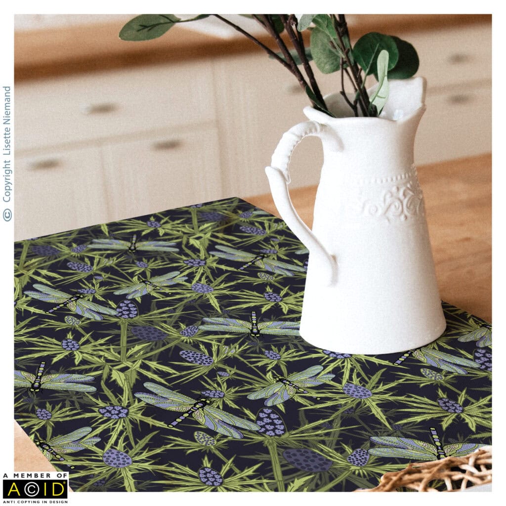 Design by Lisette Niemand called Thistle Be Sea Holly & Dragonflies shown on a home décor mock-up laid across a wooden table with a white ceramic jug placed on top. The repeat pattern features sea holly stems and dragonflies in soft honeydew green, lilac purple, cotton candy and sky blue tones on a dark lilac background.